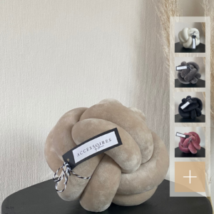 Handgefertigtes Knotenkissen in Größe M aus Samt in beige, abgebildet auf einem schwarzen Tisch. Auf der rechten Seite werden kleine Icons mit den verfügbaren Farben des Kissens angezeigt: cremeweiß, grau, schwarz und rosa.