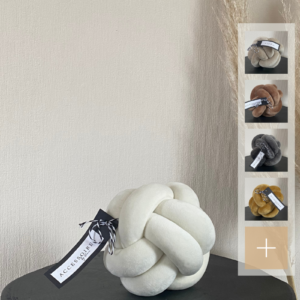Handgefertigtes Knotenkissen in Größe S aus Samt in cremeweiß, abgebildet auf einem schwarzen Tisch. Auf der rechten Seite werden kleine Icons mit den verfügbaren Farben des Kissens angezeigt: hellbeige, dunkelbeige, grau und goldgelb.