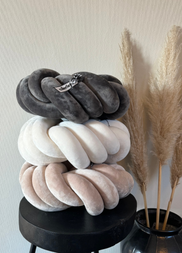 Drei gestapelte Swirl-Knotenkissen in den Farben cremeweiß, hellbeige und grau. Aus weichem Samt. Sie liegen auf einem kleinen Tisch.
