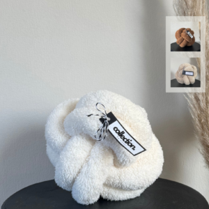 Knotenkissen aus weichem Teddystoff in cremeweiß Gr. M auf einem schwarzen Tisch. Daneben werden die weiteren Farben in einem Kästchen gezeigt. Diese sind Karamell und hellbeige
